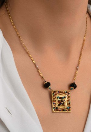 Collier chaîne en or avec un pendentif carré orné de pierres multicolores et de perles noires, porté avec une chemise blanche.