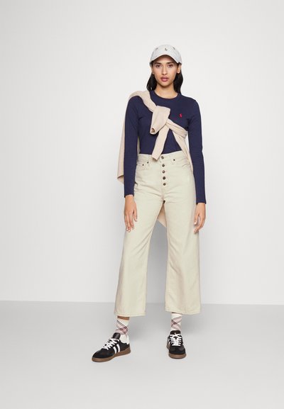 Granatowa koszula z długim rękawem z małym logo, beżowe spodnie o wysokim stanie i szerokich nogawkach z przednimi guzikami, szara czapka oraz czarne trampki z białymi sznurówkami.