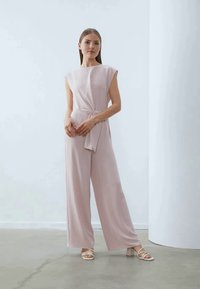 Lys rosa ermeløs jumpsuit med knyttet midje og vid bendesign. Har en jevn tekstur og en uthevet minimalistisk silhuett.