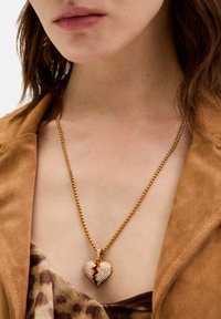 Collana in oro con pendente a forma di cuore spezzato, decorato con strass trasparenti e un accento rosso, appoggiata su una maglietta beige.
