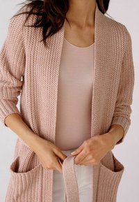 Cárdigan rosa de punto con apertura frontal, tejido texturizado y dos bolsillos delanteros. Lleva una camiseta rosa claro debajo, mostrando detalle en la manga.