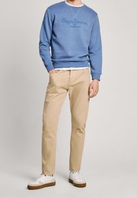 Sudadera azul claro con cuello redondo y detalle de logo, combinada con pantalones beiges y zapatillas blancas con suelas marrones.