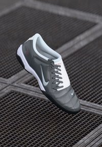 Os sapatos de futebol da Nike apresentam um cabedal sintético cinza com a ponta e a área de atacadores brancas, superfície texturizada e uma sola de borracha preta.