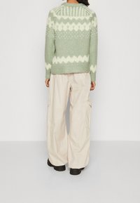 Zielony sweter dzianinowy z białym wzorem w zygzak, z długimi rękawami i okrągłym dekoltem, w połączeniu z jasnobeżowymi spodniami cargo i czarnymi botkami.