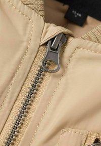 Tessuto giacca beige con cerniera metallica. Il primo piano mostra la texture liscia, le cuciture visibili e il dettaglio del tirante della cerniera.