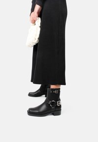 Botas negras de cuero hasta el tobillo con una correa y un detalle de hebilla plateada. Las botas tienen una puntera redondeada y un tacón bajo en bloque.