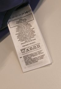 Bijela etiketa za njegu tkanine priložena tamnoplavom odjevnom predmetu. Tekst uključuje upute za pranje i višelingvalne podatke o proizvodnji.