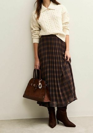 Femme portant un pull crème en maille torsadée et une jupe midi à carreaux marron, tenant un sac à main en daim marron et portant des bottines marron à talons.