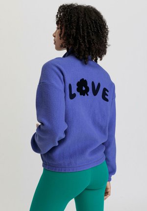 Veste en polaire de couleur violette vive avec le texte "LOVE" en noir et un détail florale au dos. Associée à un legging vert. Texture douce, coupe décontractée.