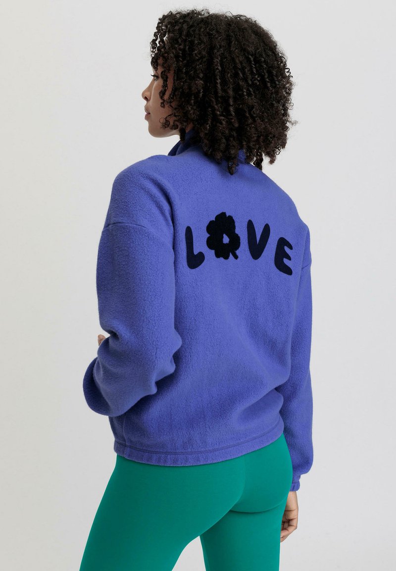 Fleecejack in een levendige paarse kleur met zwarte "LOVE" tekst en bloemenmotief op de achterkant. Gecombineerd met groene legging. Zachte textuur, relaxte pasvorm.