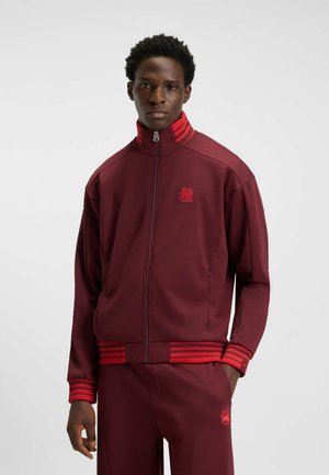 Maroon zip-up jacket with red stripes on the collar and cuffs. Features a smooth texture and subtle branding on the chest.
