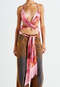 Top avvolgente con motivo floreale rosa e spalline sottili, abbinato a pantaloni marroni con una sciarpa frangiata rosa legata in vita.
