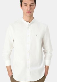 Camicia bianca con bottoni in cotone, con colletto alla coreana, maniche lunghe e un piccolo logo blu sul petto. Texture liscia.
