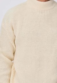 Pull en tricot beige avec un col montant, présentant un motif texturé et une coupe décontractée. Le matériau doux offre chaleur et confort.