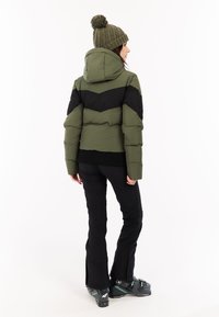 Vrouw in een groen en zwart gewatteerd jack, olijfgroene gebreide muts met pompon, zwarte broek en grijze skiboots, staand met de rug naar de camera gekeerd.