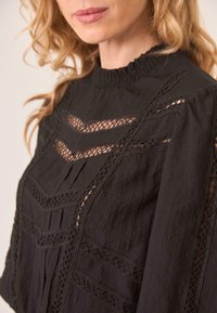 Blusa negra con tejido texturizado, inserciones de encaje y detalles plisados. Diseño de cuello alto con fruncido a lo largo del collar.