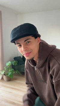 Ein junger Mann mit schwarzer Kappe und braunem Sweatshirt mit Reißverschluss, lockigem Haar, lächelt in einem hellen Innenraum mit einer Pflanze.