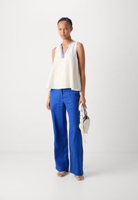 Woman wearing sleeveless white blouse with blue embroidery, bright blue wide-leg pants, white sandals, and holding a white handbag.