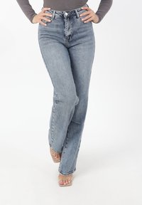 Kobieta ubrana w wysokie, jasnoniebieskie dżinsy i beżowe, odkryte na palcach szpilki, stojąca ze skrzyżowanymi nogami i rękami na biodrach na białym tle.