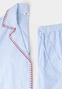Haut de pyjama rayé bleu clair et blanc avec détail de couture rouge et pantalon à cordon assorti sur fond blanc.