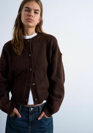 Jonge vrouw die een donkerbruine gebreide cardigan met knopen draagt over een witte top en een blauwe spijkerbroek, staand met handen in de zakken tegen een effen achtergrond.