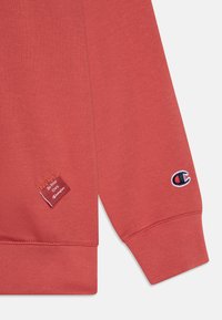 Klassisk crewneck sweatshirt i korallrød, med ribbestrikket mansjetter og kant, samt et lite stoffmerke og brodert logo på ermet.
