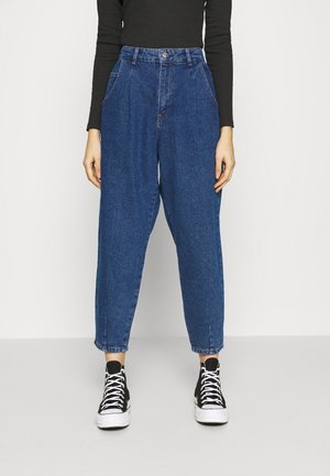 Mujer con vaqueros de mezclilla azules de tiro alto y corte holgado, combinados con zapatillas negras de caña alta y una camiseta ajustada de manga larga negra.