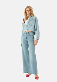 Chaqueta de mezclilla azul claro recortada con dos bolsillos en el pecho, combinada con jeans de pierna ancha de cintura alta azul claro y tacones rojos brillantes.