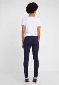 Femme aux cheveux bouclés portant une chemise blanche à manches courtes, un pantalon slim bleu marine et des sandales noires à talons hauts, debout de dos.