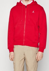 Man in een rode zip-up hoodie met een wit logo op de borst en beige broek met een klein logo nabij de zak.