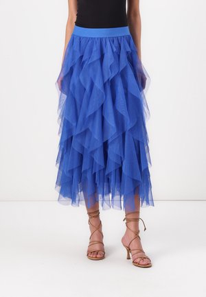 Jupe midi en tulle bleu superposée, associée à un top noir et des sandales marron à talons hauts et à brides, portée par un mannequin sur fond blanc.