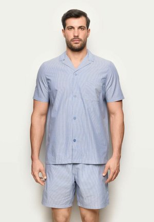 Mężczyzna w jasnoniebieskiej koszuli na krótki rękaw zapinanej na guziki i pasujących do niej szortach, stojący na tle jednolitego jasnego tła.