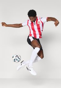 Camisa de futebol com riscas vermelhas e brancas, calções pretos, bola de futebol e chuteiras brancas. Jogador está no meio de um salto, demonstrando ação atlética.