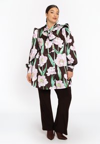 Langärmlige Bluse aus braunem Satin mit rosa floralem Druck, mit einem gebundenen Kragen-Detail, kombiniert mit dunkelbraunen, eng anliegenden Hosen und spitzen Absätzen.