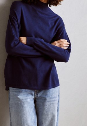 Person iført en navyblå rullekrave med lange ærmer krydset, kombineret med lyseblå denimjeans mod en ensfarvet lys baggrund.