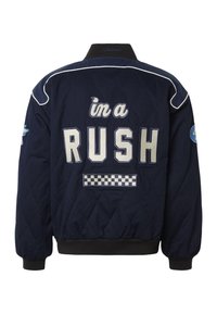 Veste matelassée bleu marine avec passepoil blanc, patch à carreaux et grand texte « i'm a RUSH » dans le dos. Col et poignets noirs.