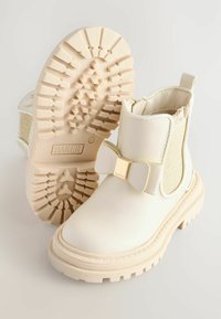 Weiße Knöchelstiefel mit einer glänzenden Oberfläche, elastischen Seitenteilen, einer goldakzentuierten Schleife und einer strukturierten Gummisohle mit einem Profilmuster.