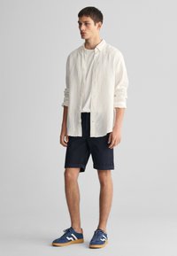 Camisa blanca de manga larga, pantalones cortos de color marino y zapatillas de ante azul con detalles en blanco. El atuendo presenta un corte relajado y un diseño casual.