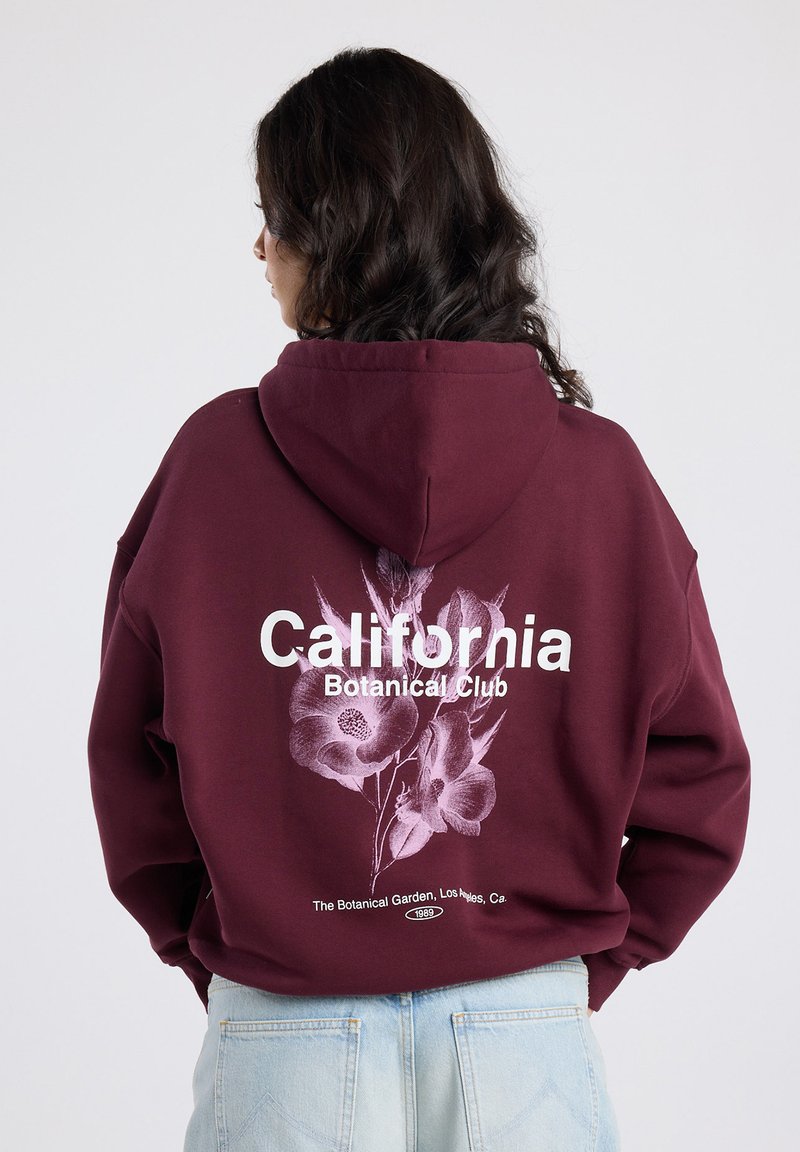 Person with dark hair wears a maroon hoodie showing "California Botanical Club" and floral graphic over light blue jeans, facing away.