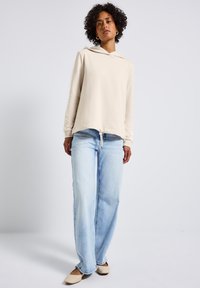 Felpa beige con cappuccio e orlo arrotondato, abbinata a jeans a gamba larga blu chiaro e ballerine beige. Presenta un cordino e polsini a costine.