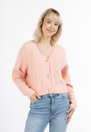 Růžový pletený cardigan z měkkého materiálu, s výstřihem do V, ribbovaným lemem a velkými knoflíky, spárovaný se světle modrými džíny.