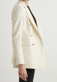 Cream-colored double-breasted blazer with gold buttons, notched lapels, side pockets, and a structured, tailored shape.
