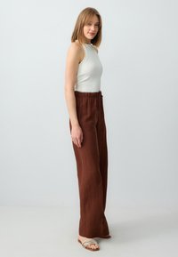 Top bianco senza maniche con bordi ondulati, abbinato a pantaloni in lino marrone a vita alta e larghi. Modella che indossa sandali beige con cinturini.