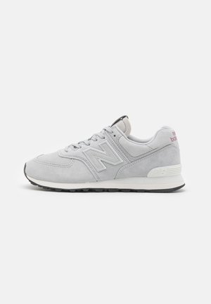 Sneaker grigia in suede con collare imbottito, parte anteriore con lacci e suola in gomma nera. Presenta un logo "N" prominente e accenti testurizzati.