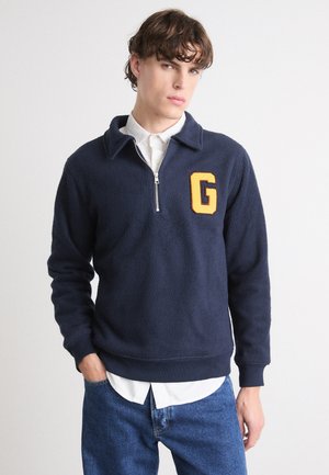 Marineblauer Fleece-Pullover mit einem weißen Reißverschlusskragen, ausgestattet mit einem gelben "G"-Emblem auf der linken Brust sowie gerippten Bündchen und Saum.