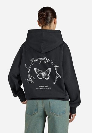 Vrouw met gevlochten knot, die een zwarte hoodie draagt met een witte vlinder en de tekst "Pegador Everyday Ambition", gecombineerd met een blauwe spijkerbroek.