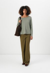 Top verde a maniche lunghe con una vestibilità comoda, abbinato a pantaloni larghi color oliva. Borsa a spalla in suede marrone e scarpe scure. Sfondo semplice.