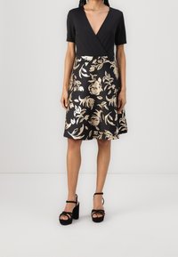 Svart klänning med djup V-ringning och korta ärmar, med ett guldblommigt mönster på en utsvängd kjol. Kombinerad med svarta plattformsandaler.