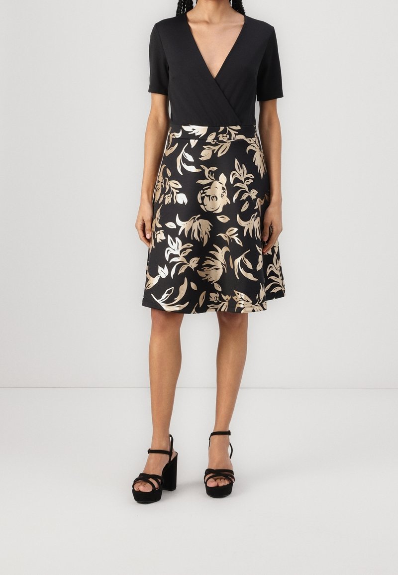 Svart klänning med djup V-ringning och korta ärmar, med ett guldblommigt mönster på en utsvängd kjol. Kombinerad med svarta plattformsandaler.