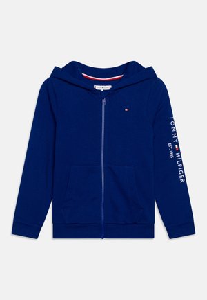 Granatowa bluza z kapturem na zamek, z przednimi kieszeniami, kapturem, małym logo na lewej piersi oraz napisem „Tommy Hilfiger EST. 1985” na prawym rękawie.